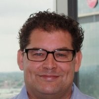 A man with curly brown hair, wearing black glasses and a light blue collared shirt, smiles in front of a window with an urban cityscape in the background.