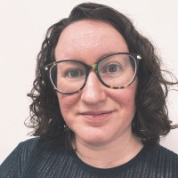 A person with fair skin, curly brown hair, and large, round spectacles is smiling softly at the camera. They are wearing a dark top and are positioned against a plain light background.