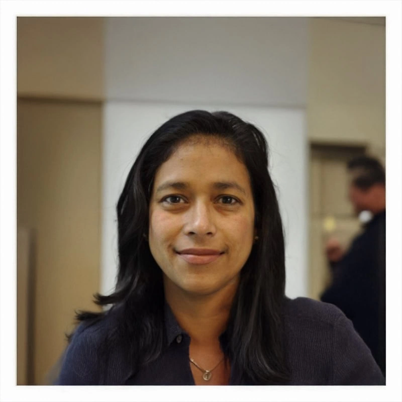 A woman with long dark hair and a slight smile faces the camera indoors. She wears a dark top and a necklace, with a blurred background and another person visible behind her.