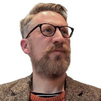 A man with light brown hair, a beard, and glasses looks slightly upward and to the side. He is wearing a brown tweed jacket over an orange jumper and striped shirt. The background is plain white.