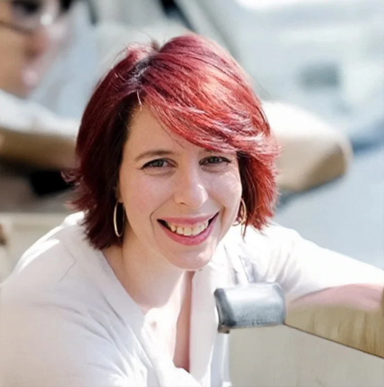 A woman with short, red hair and hoop earrings smiles whilst sitting outdoors in a car. She is wearing a white top and is holding onto a beige car door. The background is blurred.