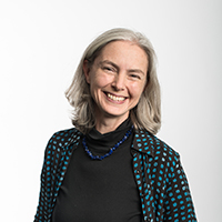 A woman with grey hair smiles at the camera. She is wearing a black top, a blue beaded necklace, and a black shirt with blue polka dots, standing against a plain white background.