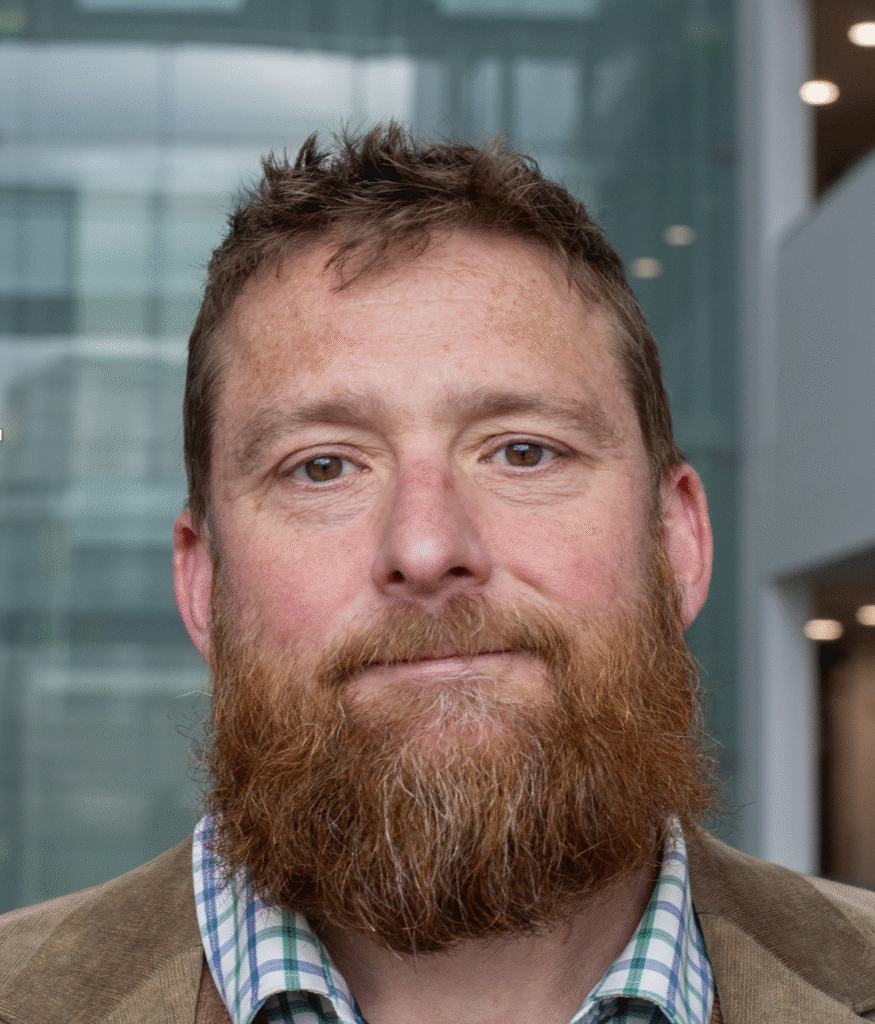 A man with a thick, reddish-brown beard and short hair is wearing a checked shirt and brown jacket, standing indoors in front of a modern glass-walled backdrop.