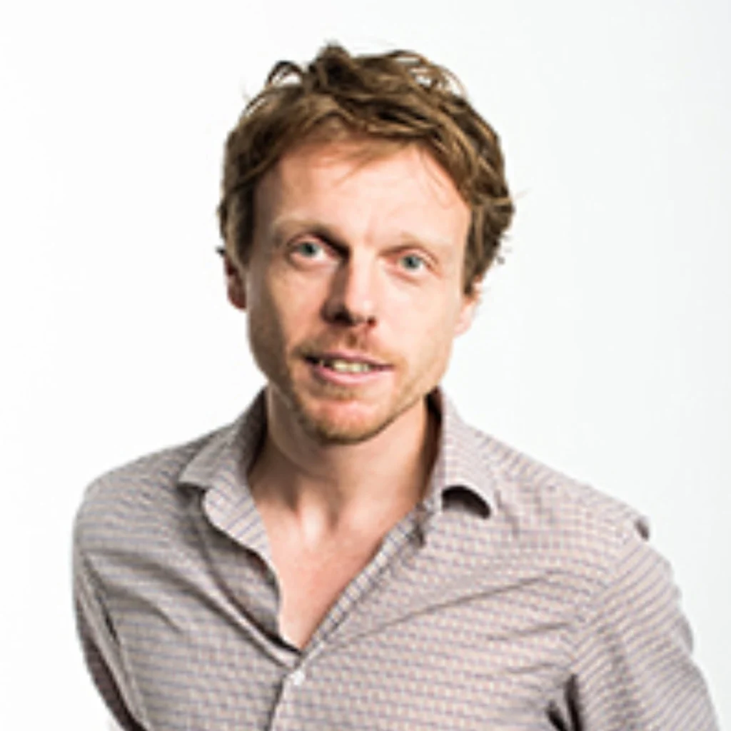A man with light brown hair wearing a light-coloured, patterned button-up shirt stands against a plain white background, looking at the camera with a slight smile.