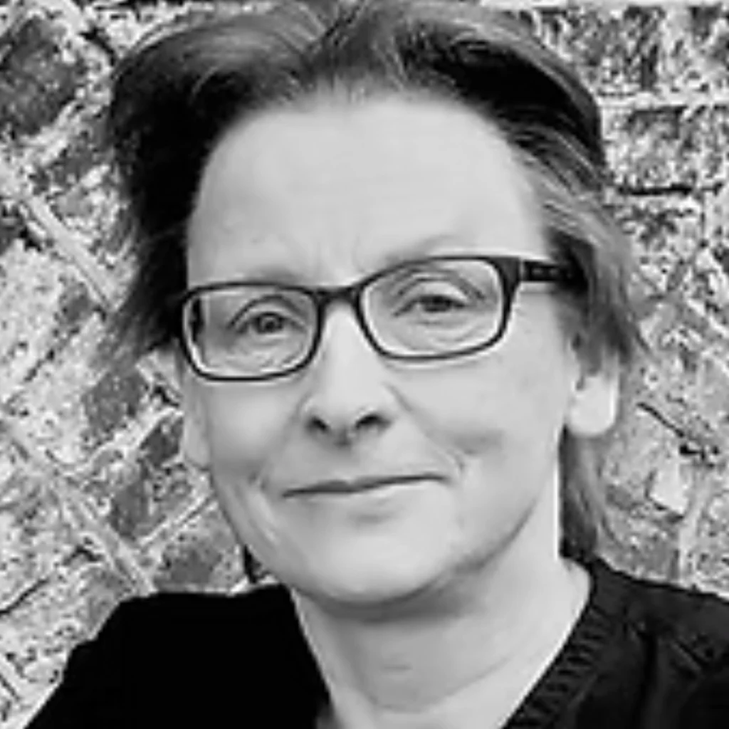 Black and white photo of a person with short hair and glasses, smiling slightly. They are standing in front of a textured stone or brick wall.