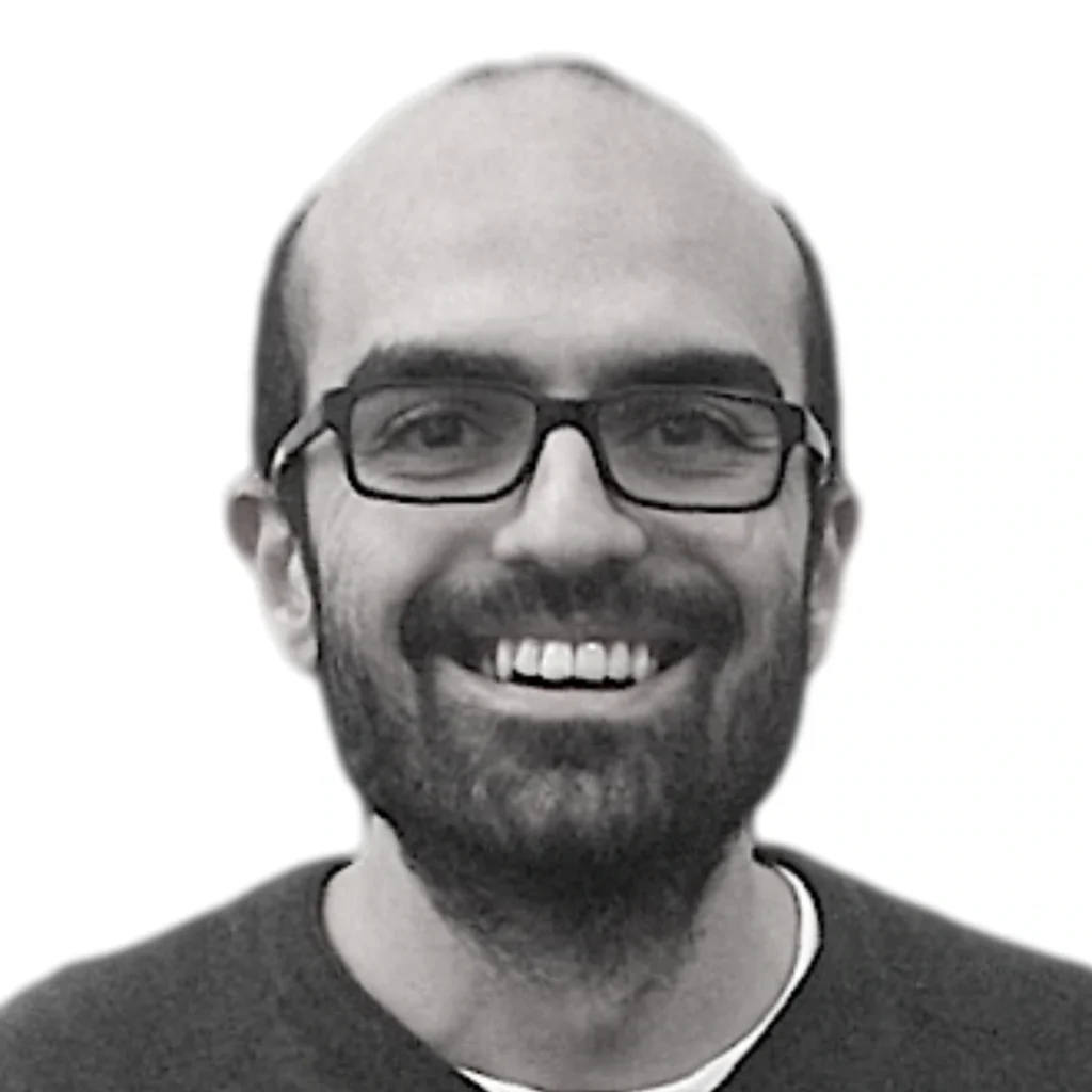 A man with a beard and glasses smiles at the camera. He has short dark hair and is wearing a dark shirt. The background is plain white.