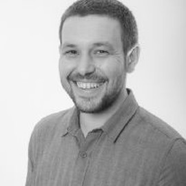 A man with short hair and a beard, smiling, is wearing a collared shirt. The photo is in black and white and has a plain, light background.